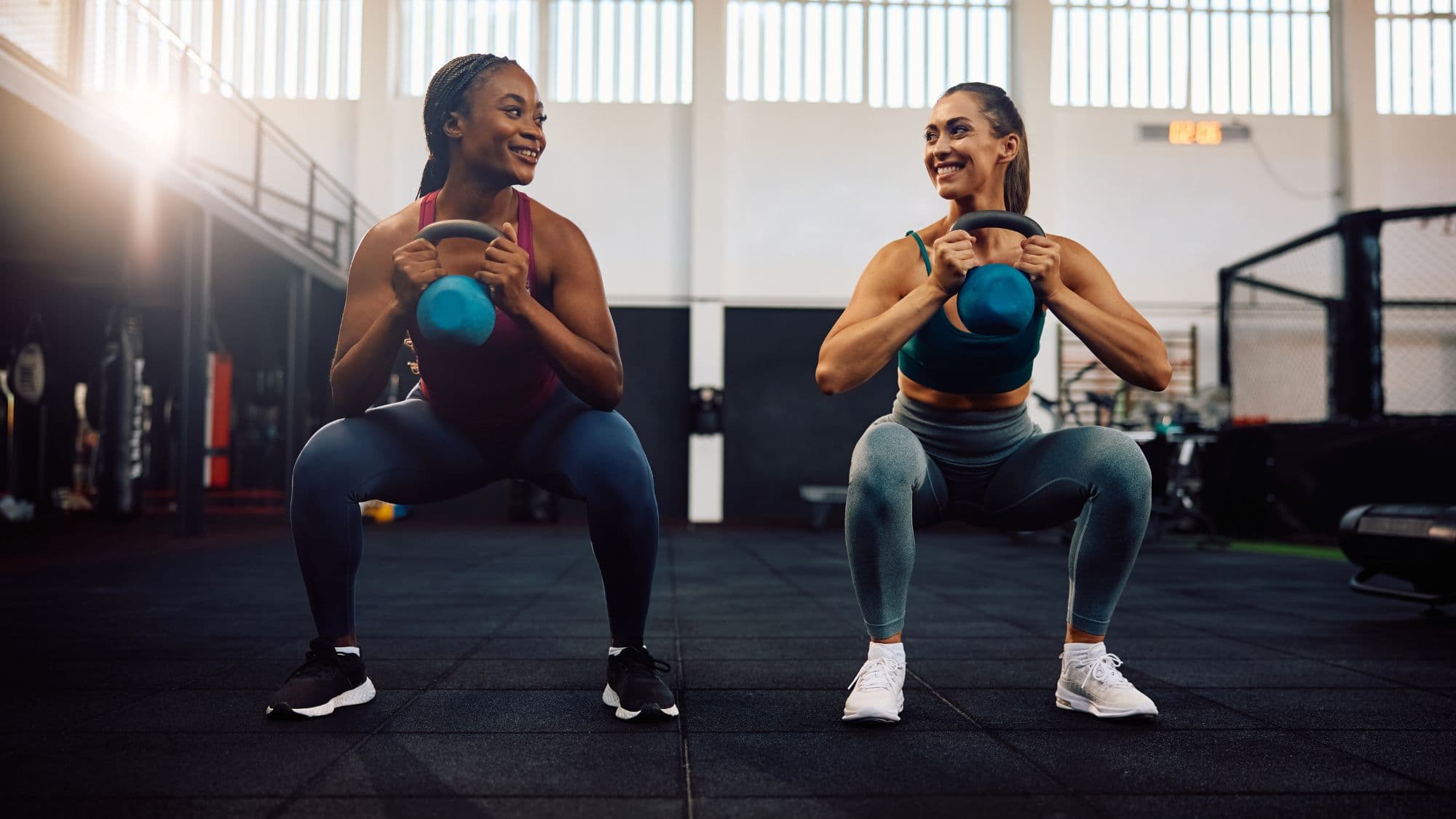 Two athletes side by side in a gym, each holding a kettlebell at chest height at the bottom of a squat